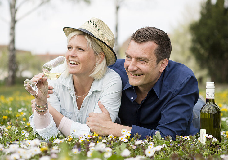Weintour Paar Ein Pärchen liegt in einer Blumenwiese mit einem Glas Wein in der Hand und lacht.