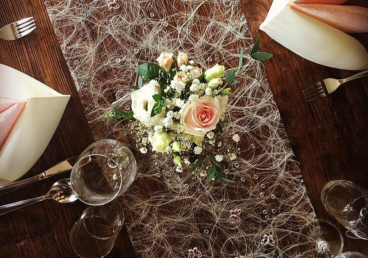 Muttertagsbuffet gedeckter Tisch In einer Vase sind geschnittene Rosen mit grünem Schleierkraut. Unter der Vase ist ein weißer Seidentischläufer. Zwei gefaltete Servietten, sowie Gabel, Messer und Gläser sind auf dem Tisch eingedeckt.