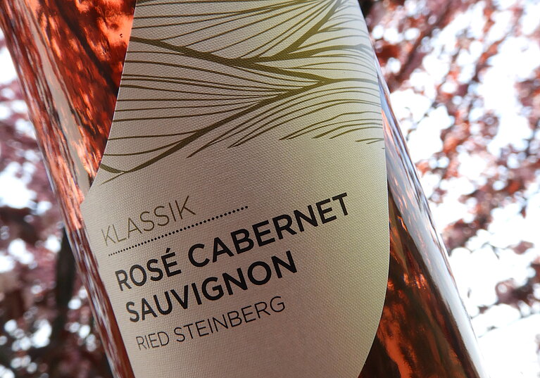 Eine Flasche Rosé Cabernet Sauvignon Klassik vom Weingut Neustifter wird vor einem rosa blühenden Baum gehalten, der seine Farbe wider spiegelt.