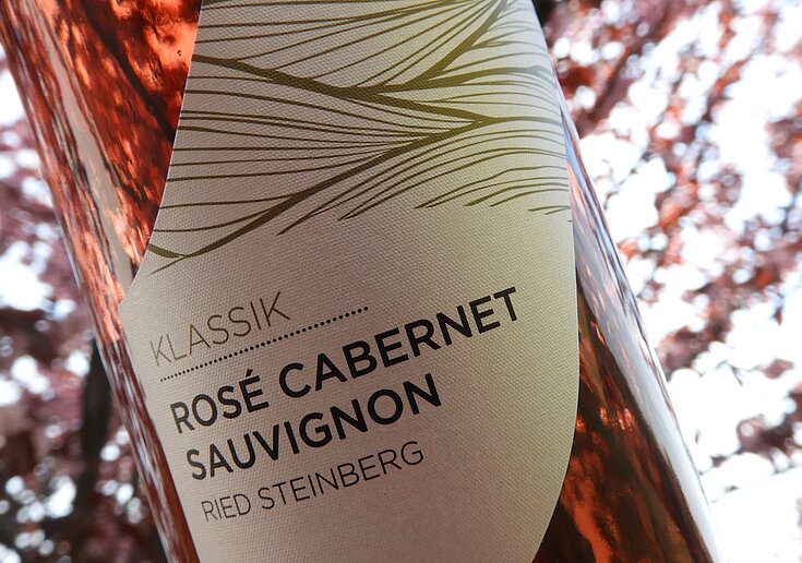 Eine Flasche Rosé Cabernet Sauvignon Klassik vom Weingut Neustifter wird vor einem rosa blühenden Baum gehalten, der seine Farbe wider spiegelt.