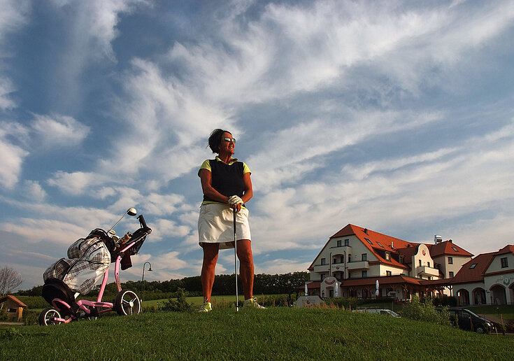 Eine Golferin steht am Golfplatz, breit den Ball abzuschlagen. Im Hintergrund sieht man das Wein.Hotel Neustifter.