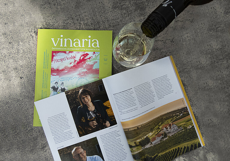 Die Zeitschrift Vinaria liegt aufgeschlagen auf dem Tisch und Zeit das Weingut Neustifter. Ein Glas Weißwein wurde dabei eingeschenkt.